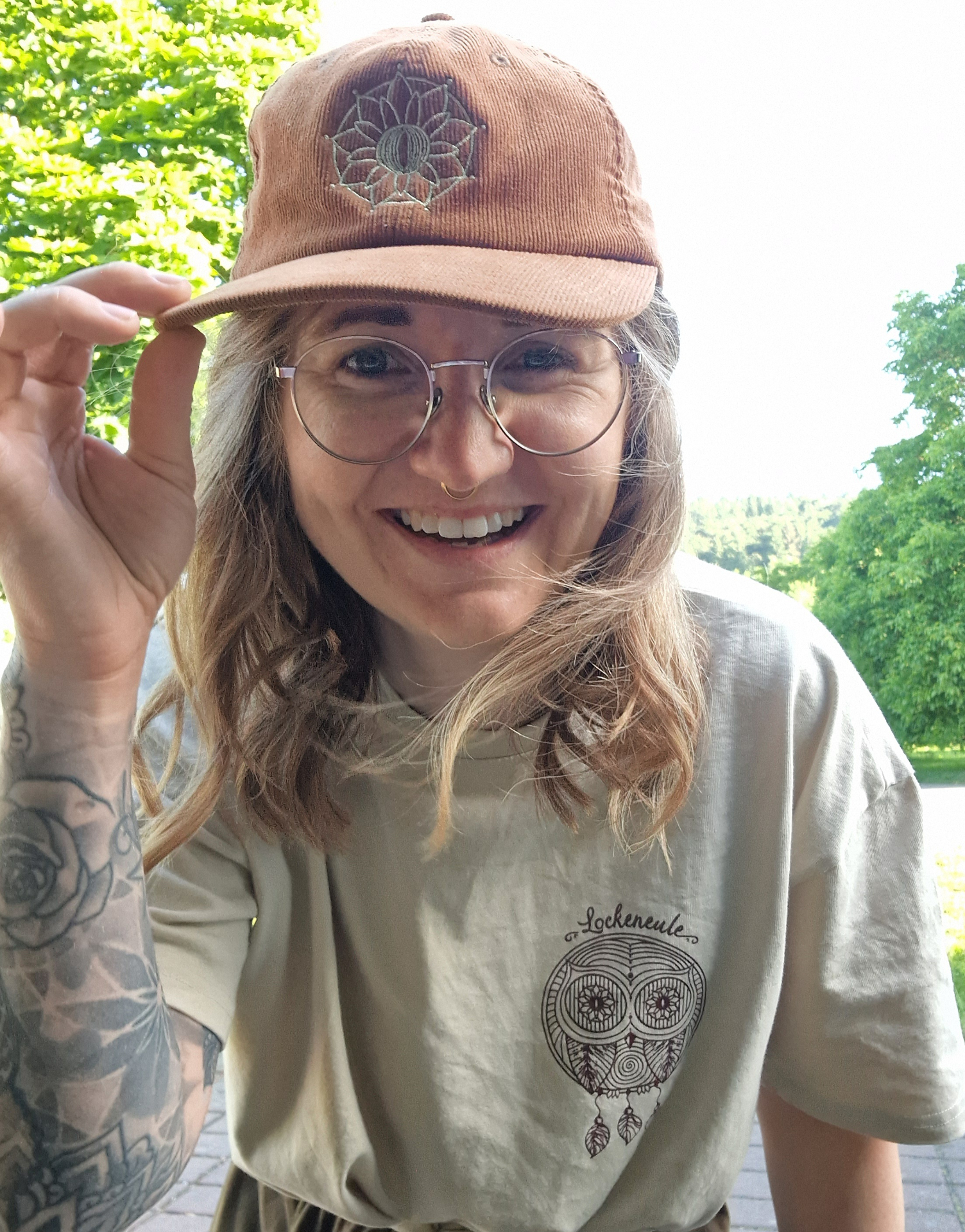 Nancy hält die Cord-Cap mit Mandala-Stickerei und trägt das Lockeneule-Shirt im Grünen